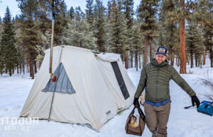 冬季露营时，一名男子在雪地里背着木柴，站在一顶热帐篷前
