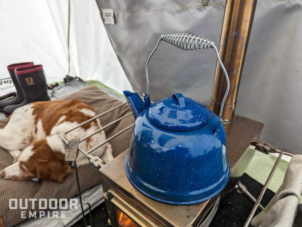 来自GSI户外的蓝色珐琅柚木水壶放在帐篷里的木炉子上，狗睡在旁边